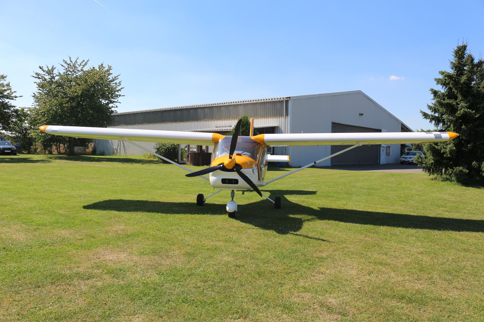 Gelb-weißes Ultraleichtflugzeug in Hochdeckerbauform auf der Wiese vor einem weißen Flugzeughangar.