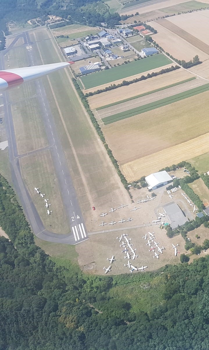 Blick aus einem Segelflugzeug auf den Flugplatz Koblenz-Winningen. Um die Hallen des Aero-Club Koblenz am unteren Bildrand stehen viele Motorflugzeuge.