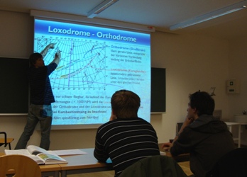 Seminarraum in dem ein Lehrer anhand einer Beamerfolie Loxodrome und Orthodrome erklärt. Schüler sitzen in den Reihen und hören zu.