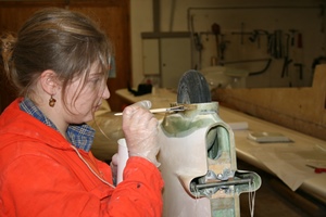 Foto einer jungen Frau, die in einer Werkstatt am Sporn eines Segelflugzeugs Glasfaser-Laminierarbeiten durchführt.