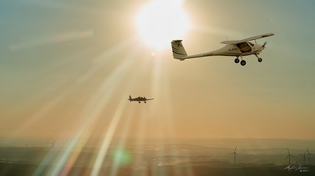 Foto eines Ultraleichtflugzeugs (Alpha Trainer) im Flug vor der Sonne, im Hintegrund ein weiteres Flugzeug.
